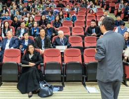 A male professor lectures in an auditorium with a crowd of students in red chairs