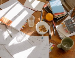 A round table is covered in open study books, a calculator and pens, class notes, sticky notes, and more textbooks. There is also an empty glass, a tea mug, and a bowl of snacks.